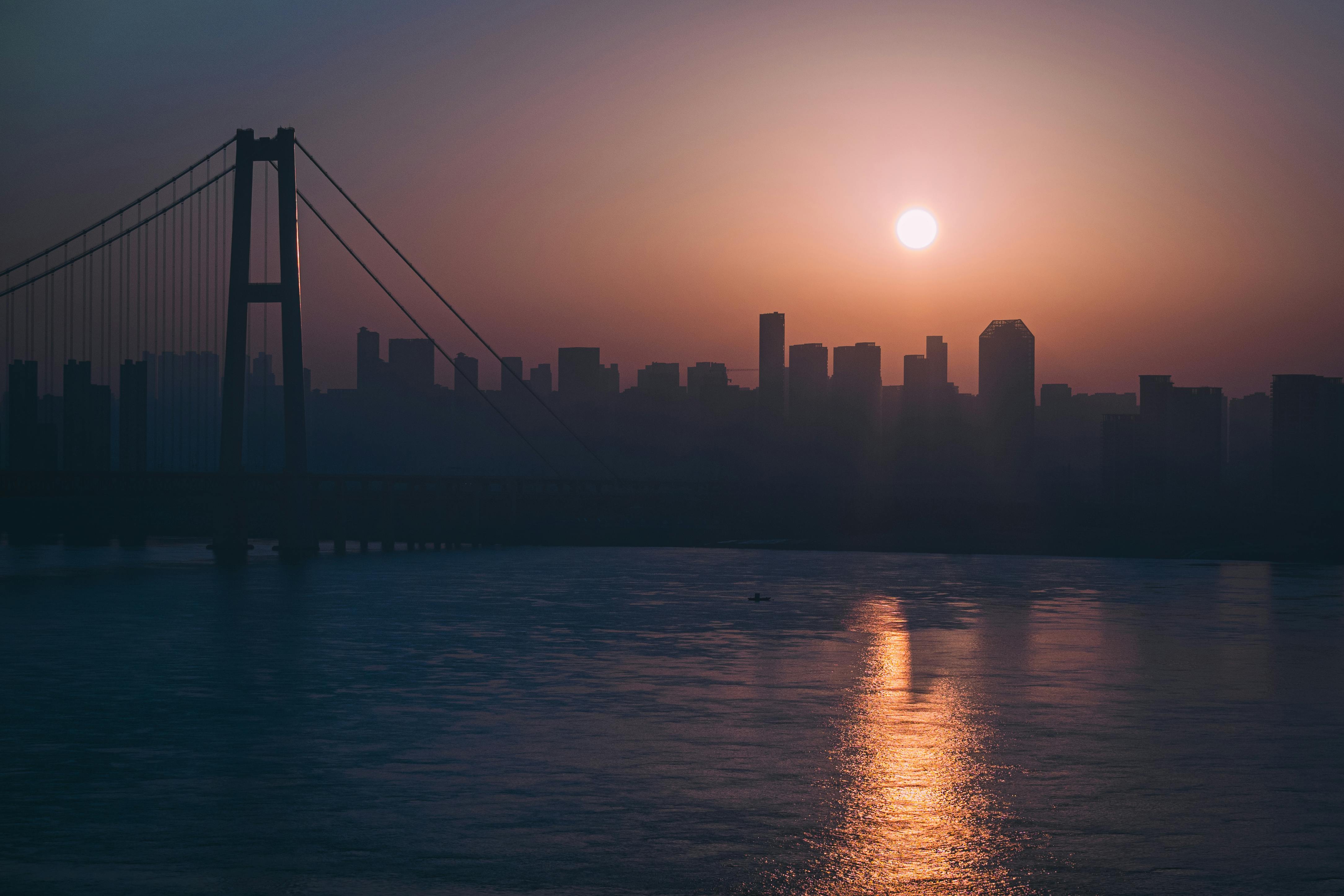 Silhouette of skyscrapers and a bridge against a stunning sunset, reflecting on the water.