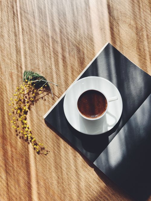 Free A top view of a coffee cup on a book with flowers on a wooden table, bathed in warm sunlight. Stock Photo