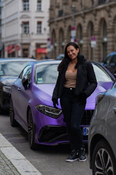 Smiling woman stands beside luxurious purple car in a vibrant city street setting.