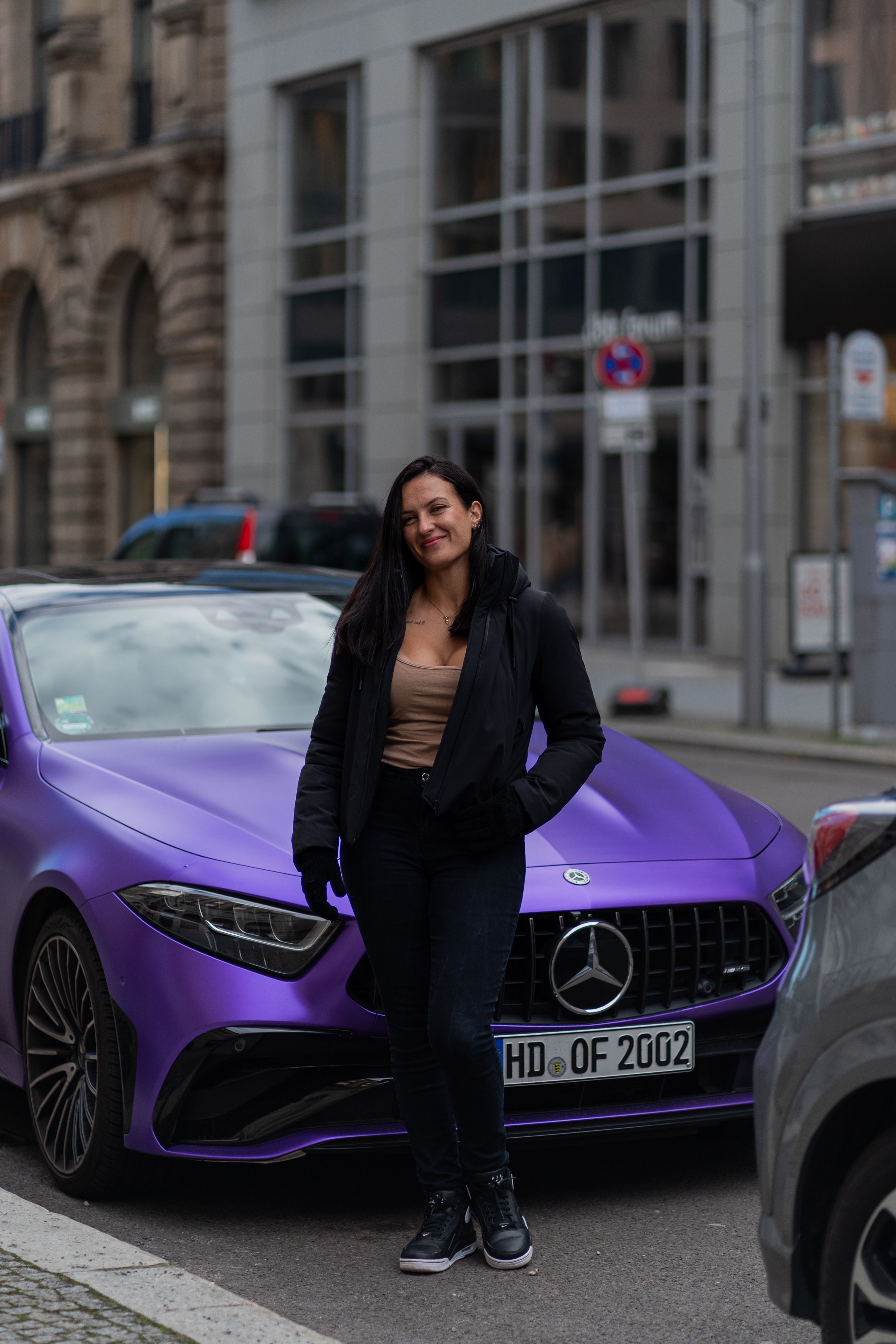Woman standing and smiling next to a stylish purple car in a vibrant city environment.