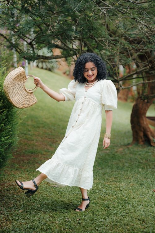 Free Woman with curly hair smiling in a park, holding a straw bag and wearing a white dress. Stock Photo