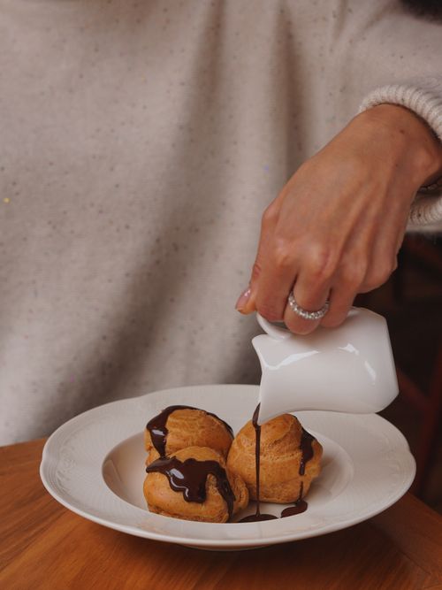 Free A woman's hand pouring chocolate sauce over profiteroles, showing dessert preparation. Stock Photo