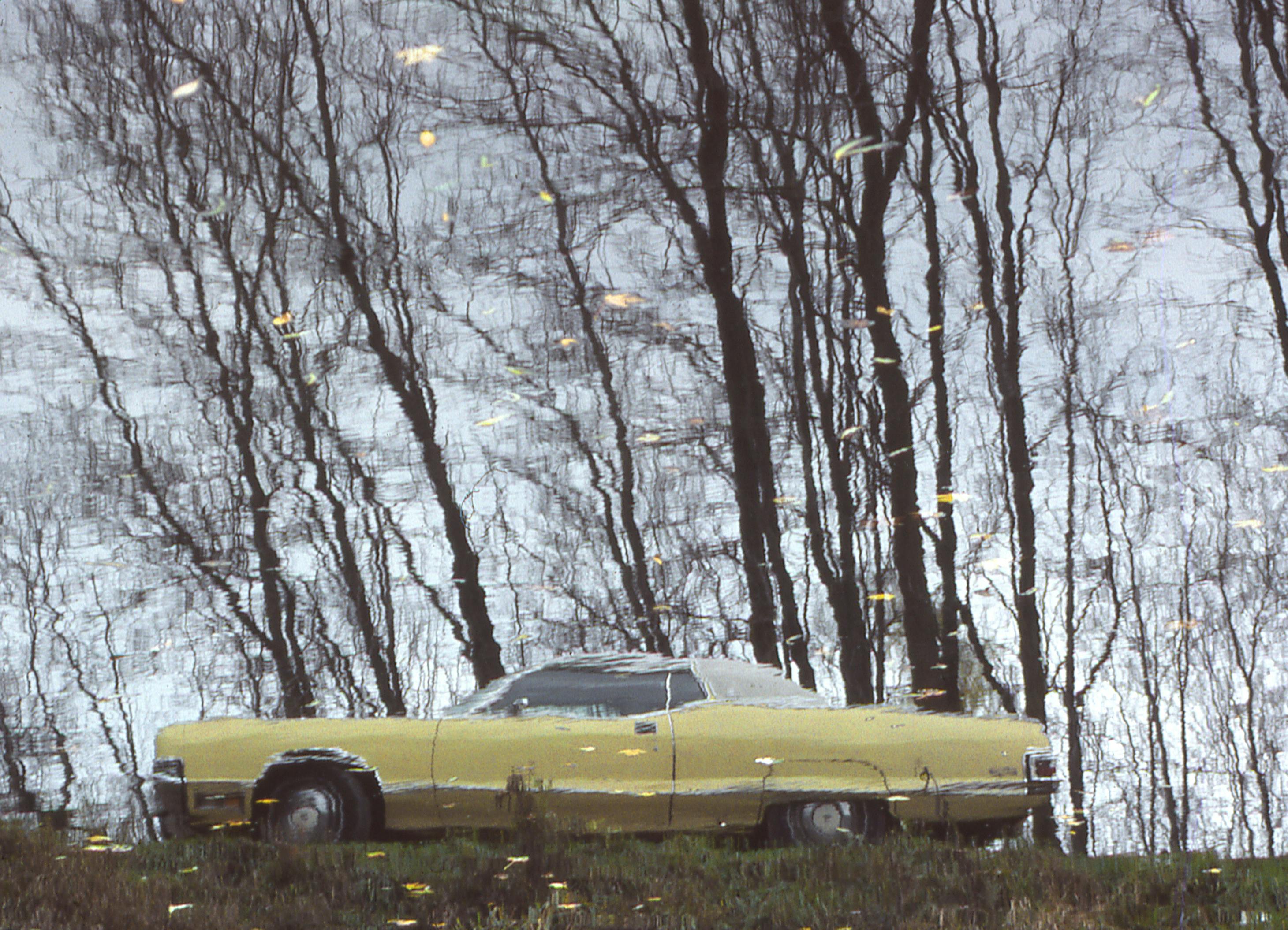 Reflection of an old-fashioned yellow car with trees in water. Artistic and serene atmosphere.