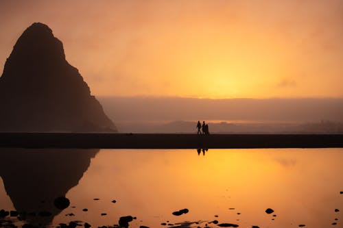 Two people stand on the beach at sunset with a mountain in the background