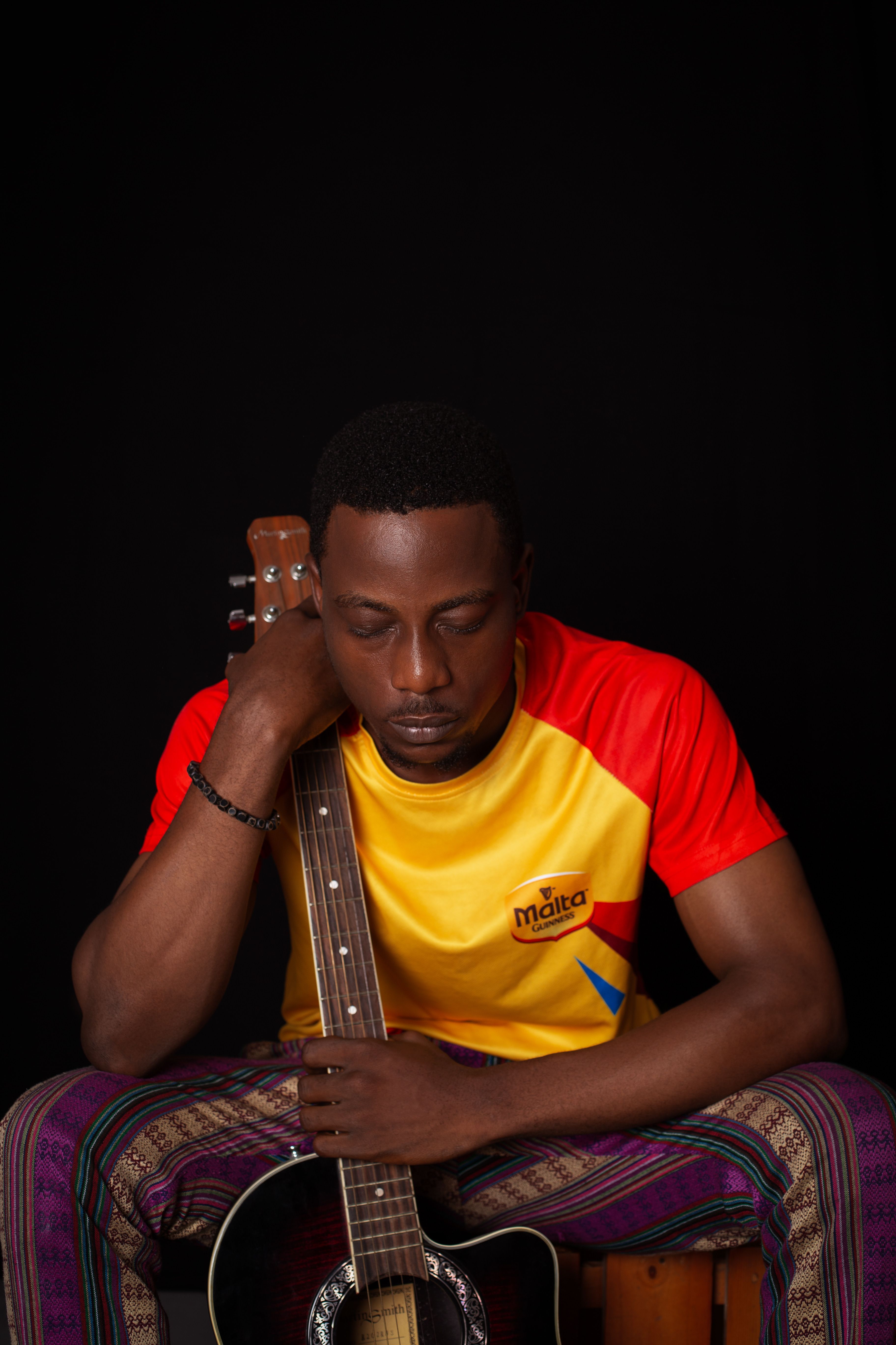 A man with eyes closed, holding a guitar, in a studio with a black background.