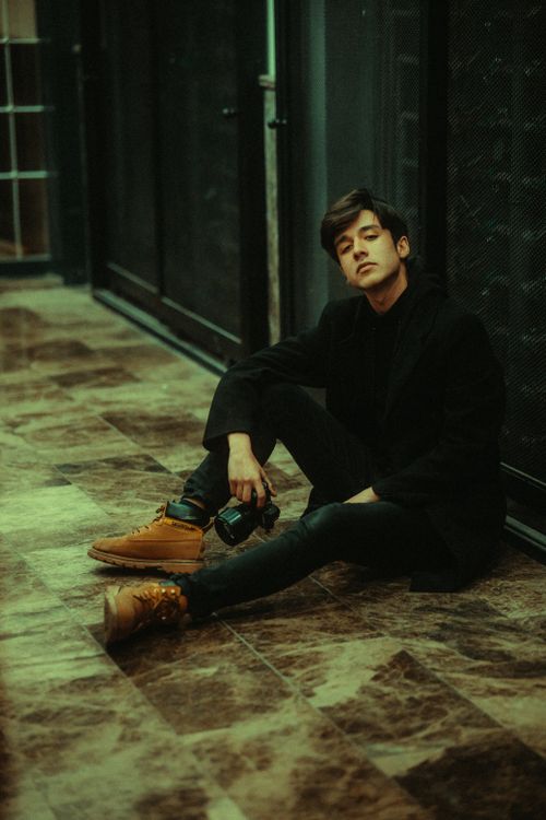 Free Stylish male model sitting on urban sidewalk in fashionable attire, holding a camera. Stock Photo