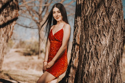 A beautiful young woman in a red dress posing near a tree