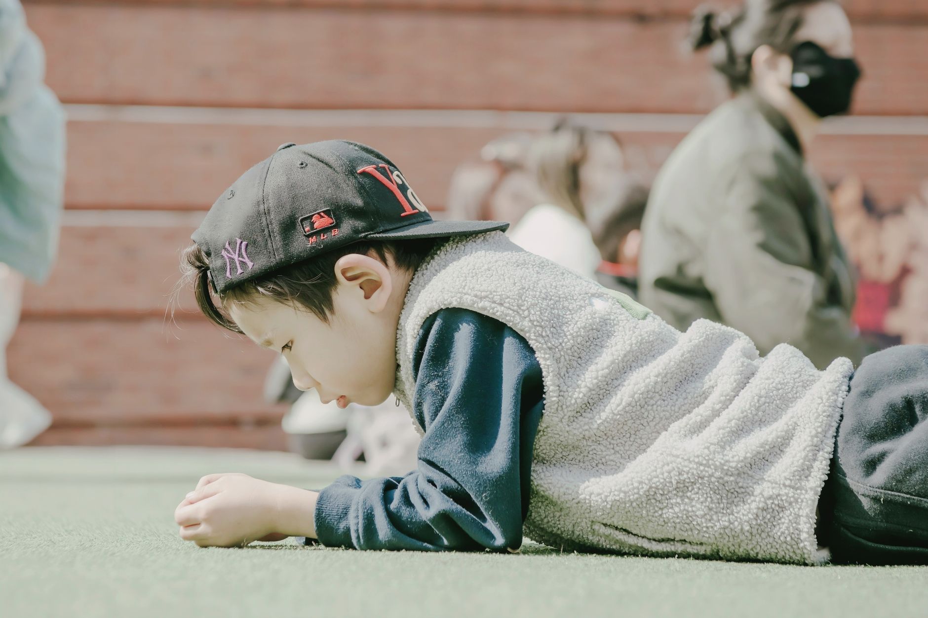A young boy in a baseball cap and sherpa vest lying on a green surface outdoors.