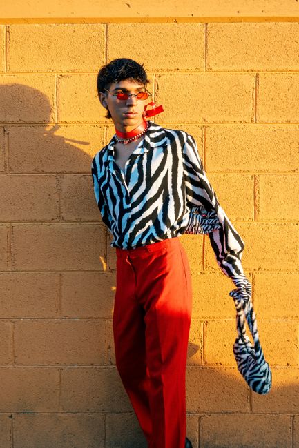 Fashionable young man in zebra print shirt and red pants leaning against yellow brick wall.