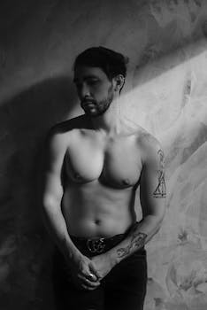 Moody black and white portrait of a topless man standing against a textured wall.