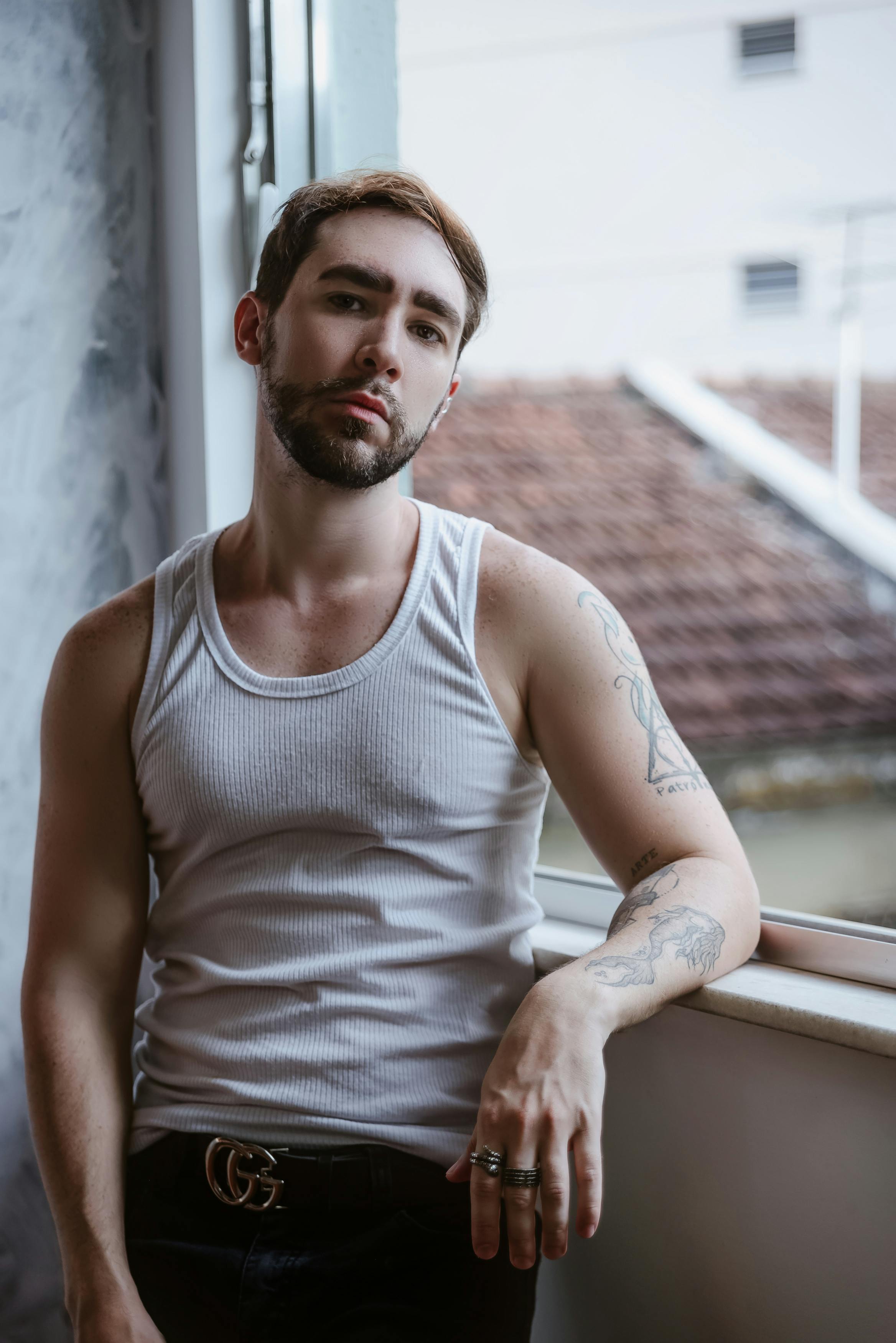Chic tattooed male model in a white tank top poses by a window in natural light.