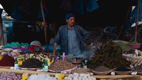 Middle-aged man in traditional attire selling spices at a vibrant outdoor market.