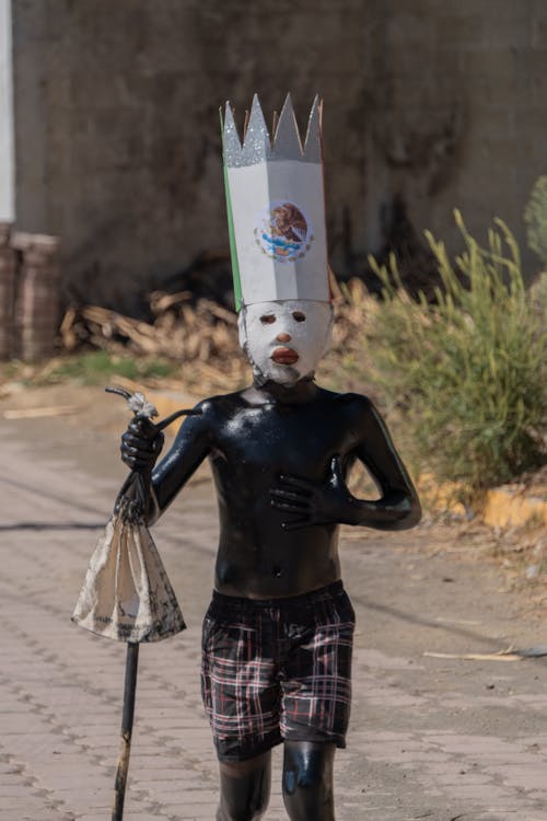 A Boy with a Mask and Headdress Walking on a Road 