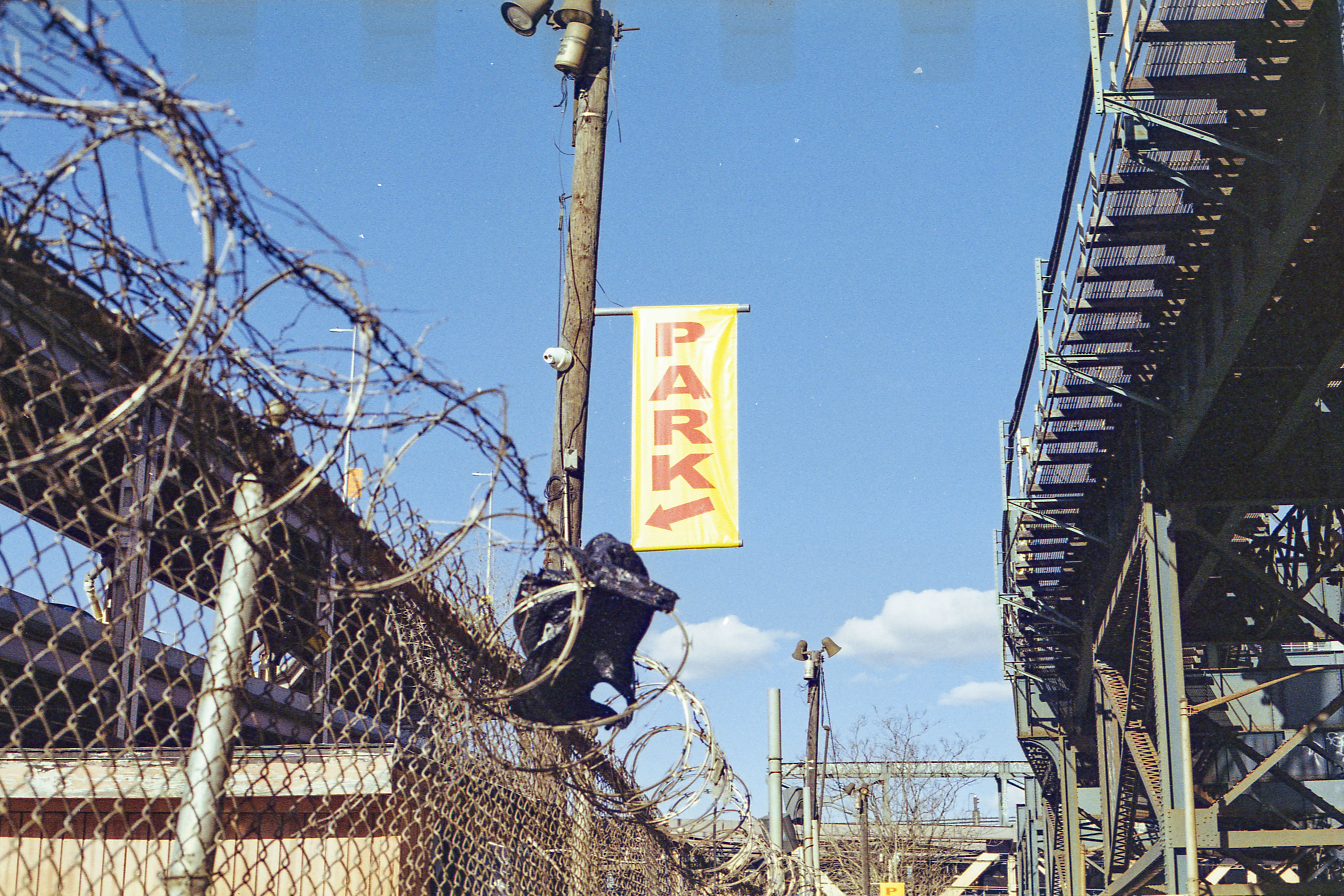 An industrial urban setting featuring barbed wire fences, metal structures, and a clear blue sky.
