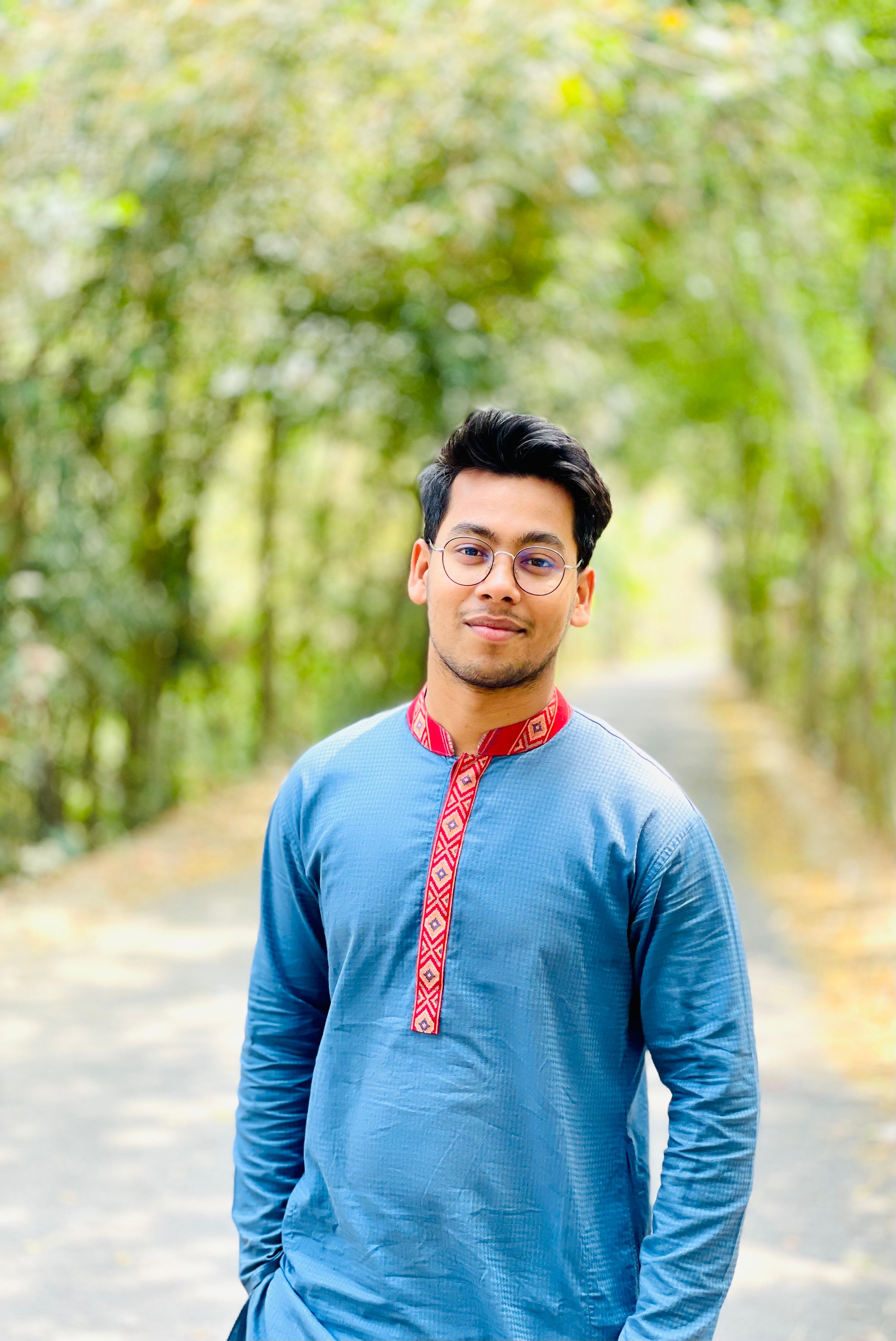 A young man in a stylish blue shirt stands relaxed in an outdoor park setting with trees.