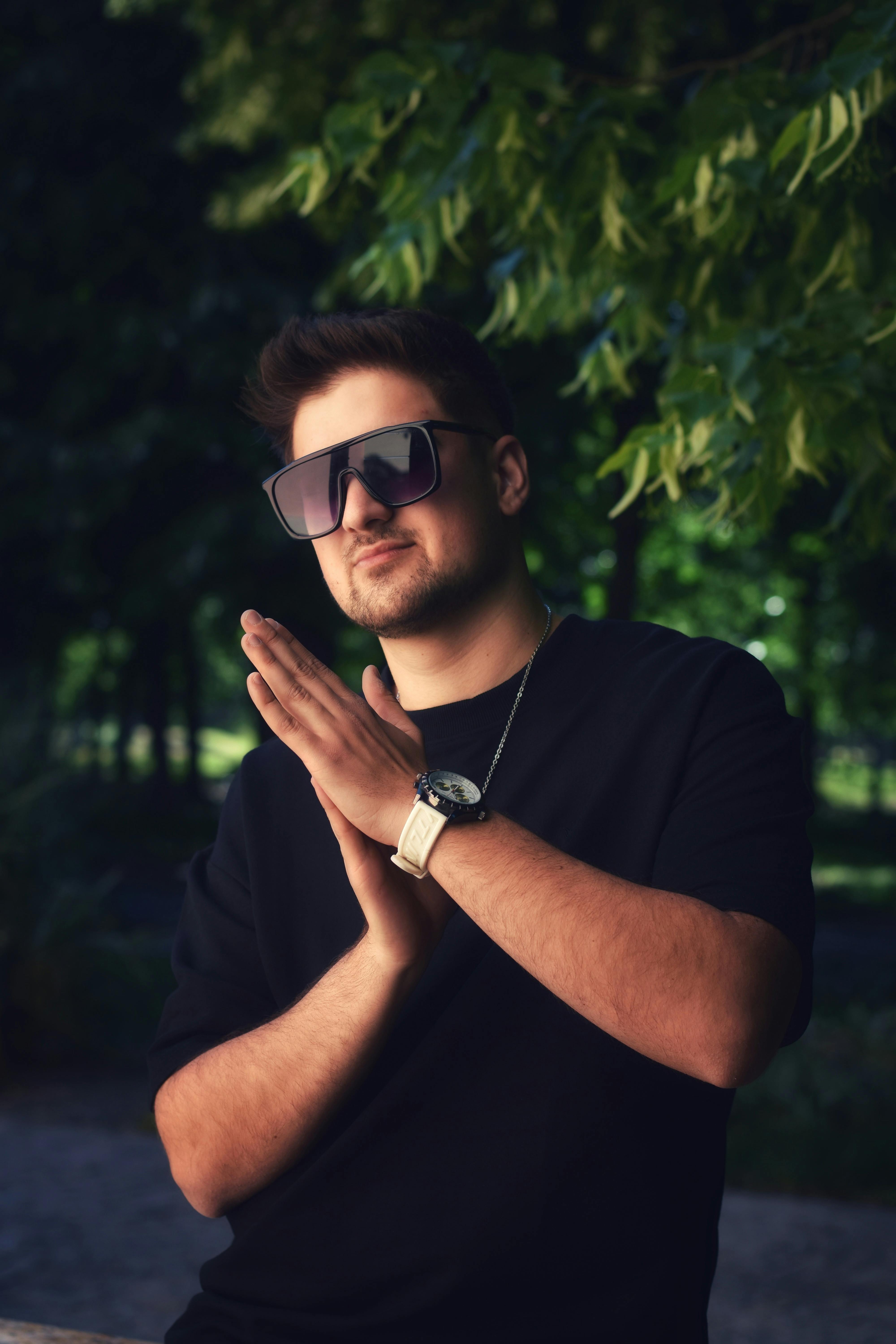 Fashionable man wearing sunglasses and black t-shirt posing outdoors with green leaves in the background.