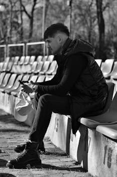 Black and white photo of a man sitting on stadium bleachers using a smartphone.