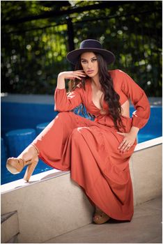 Elegant woman in a red dress with hat poses outdoors on a sunny day.
