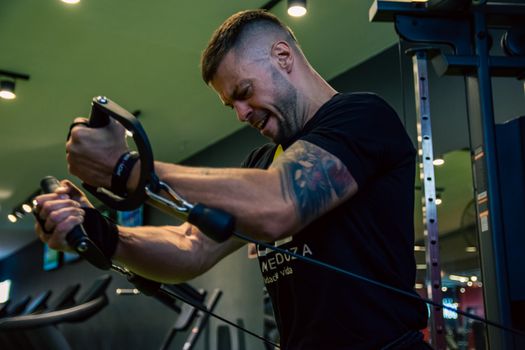 Muscular man with tattoos exercising on a cable machine in a gym, showcasing strength and determination.