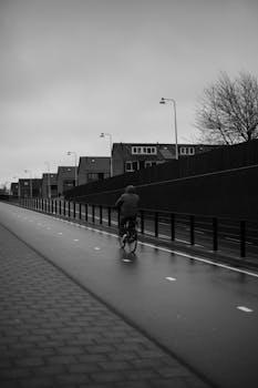 A lone cyclist rides down a deserted street on a rainy, overcast day, showcasing urban solitude.