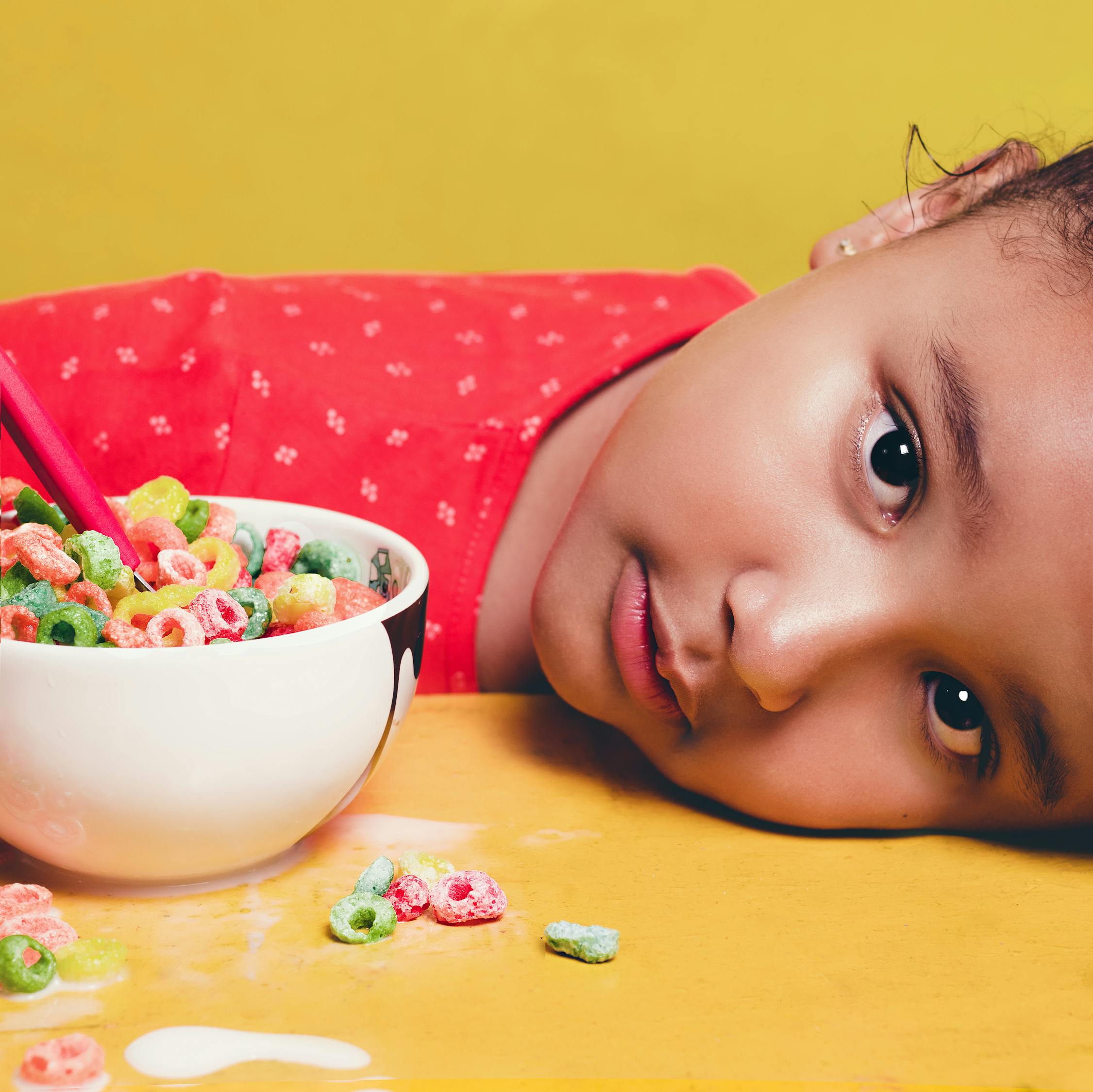 Girl Wearing Red Shirt Lying on table Beside White Bowl With Cereal