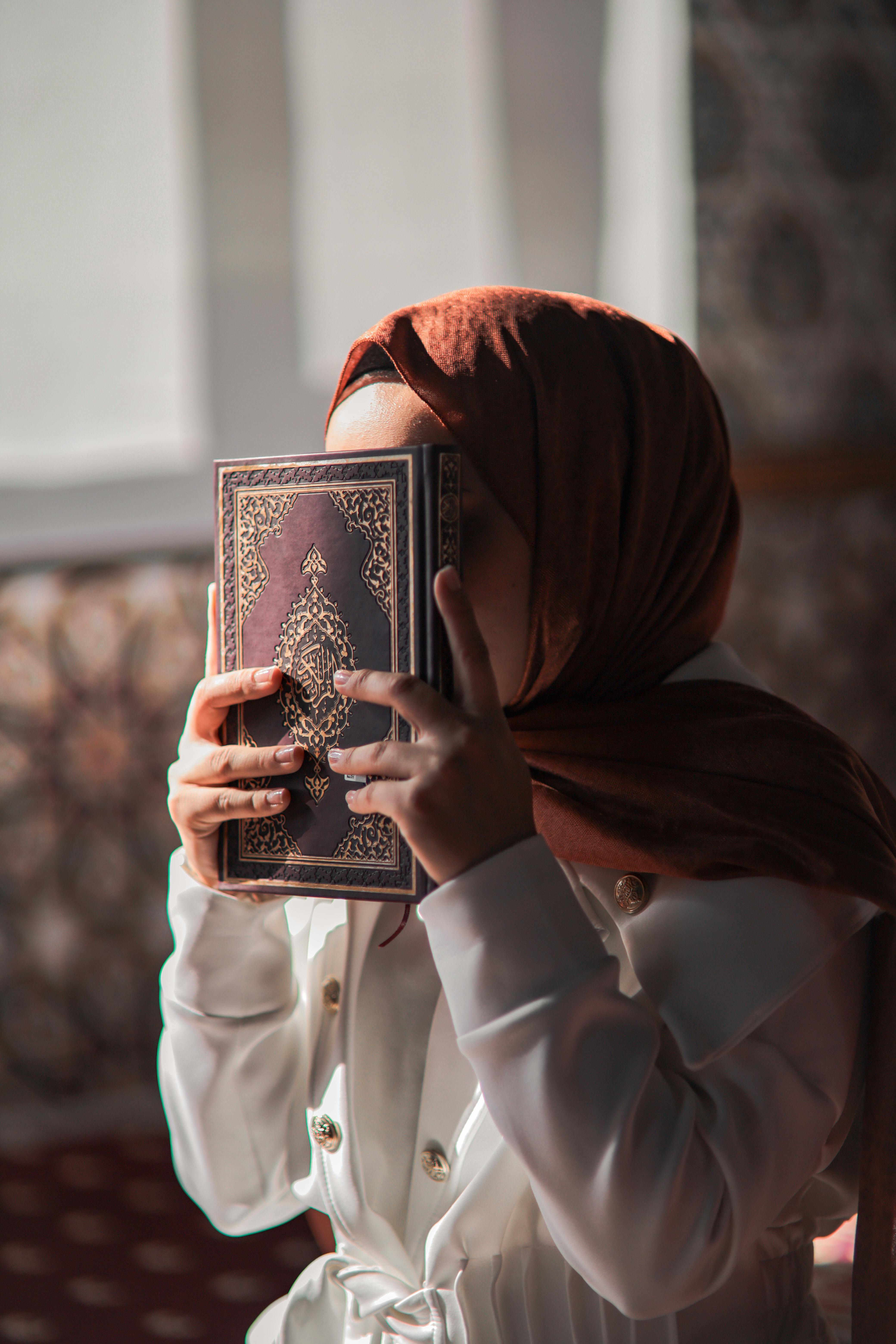 Woman in hijab holding Quran in prayer. Warm lighting, traditional setting.