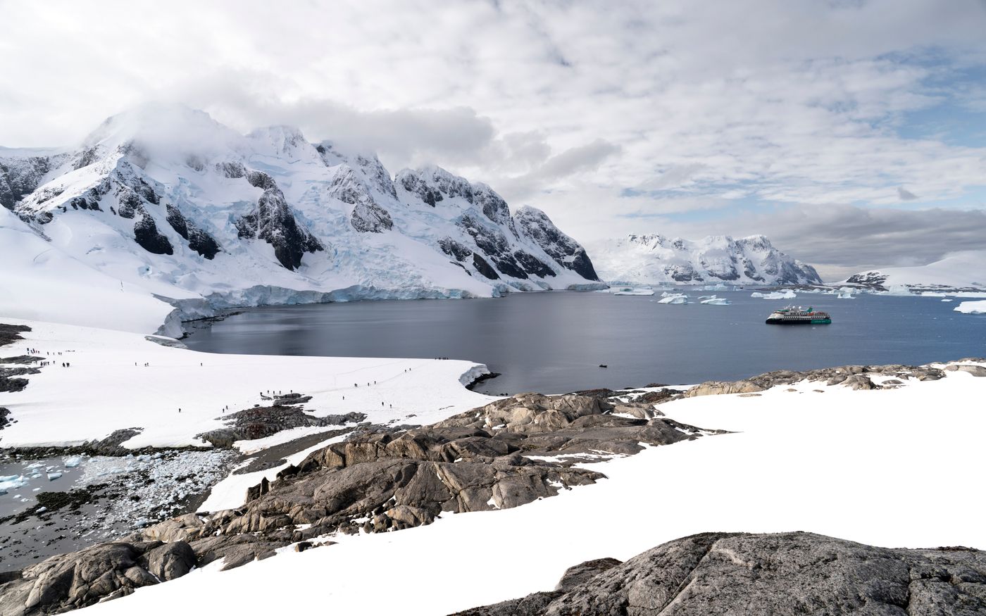 A lone vessel on calm water below snow-covered mountains in the Antarctic region