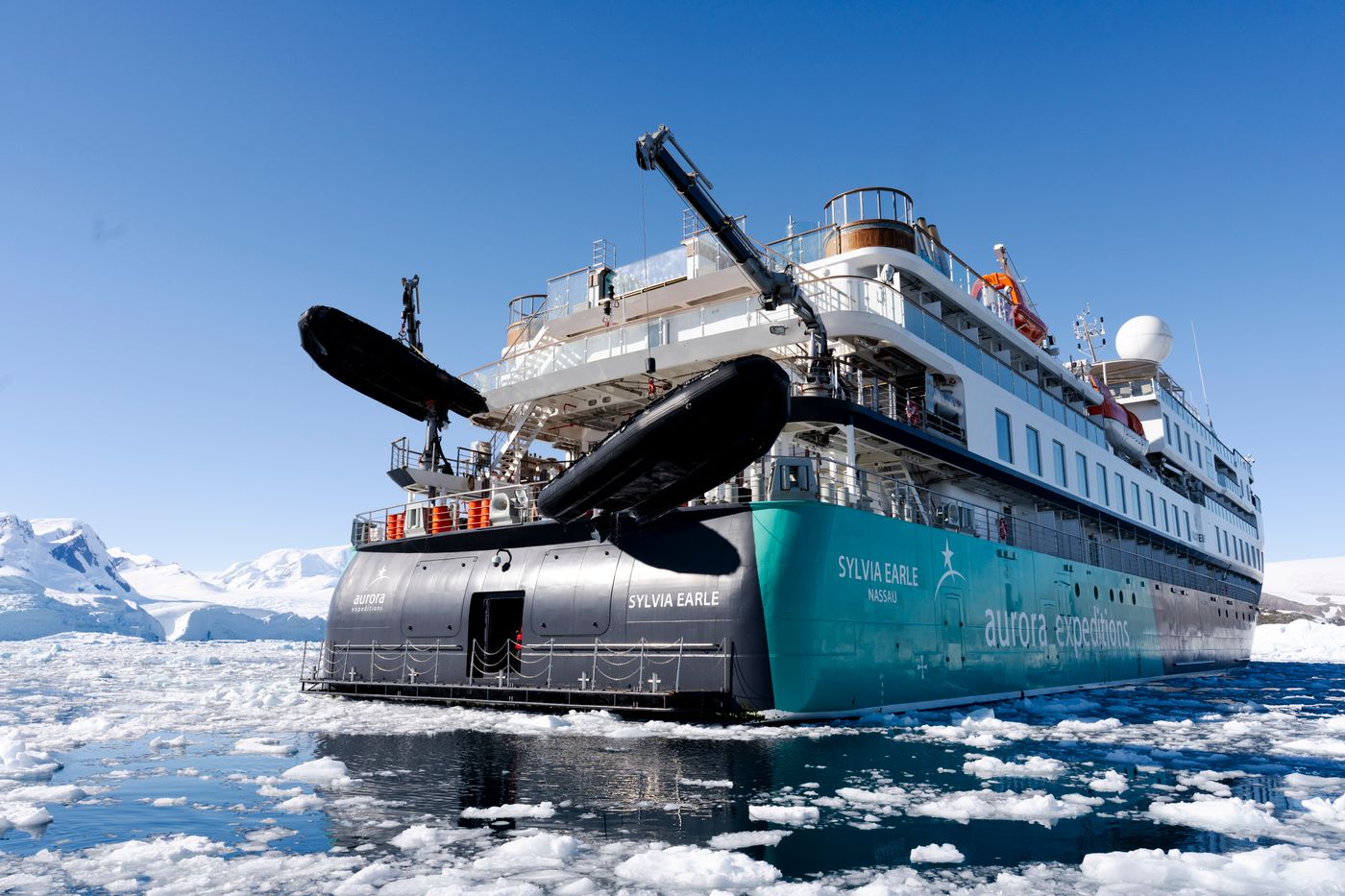 The expedition ship Sylvia Earle among icebergs in calm Arctic water under a clear sky.