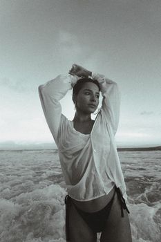 A woman poses gracefully at the beach wearing a wet shirt, blending with the ocean waves.