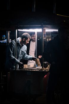 Street vendors cook food at a nighttime city market illuminated by dim lights.