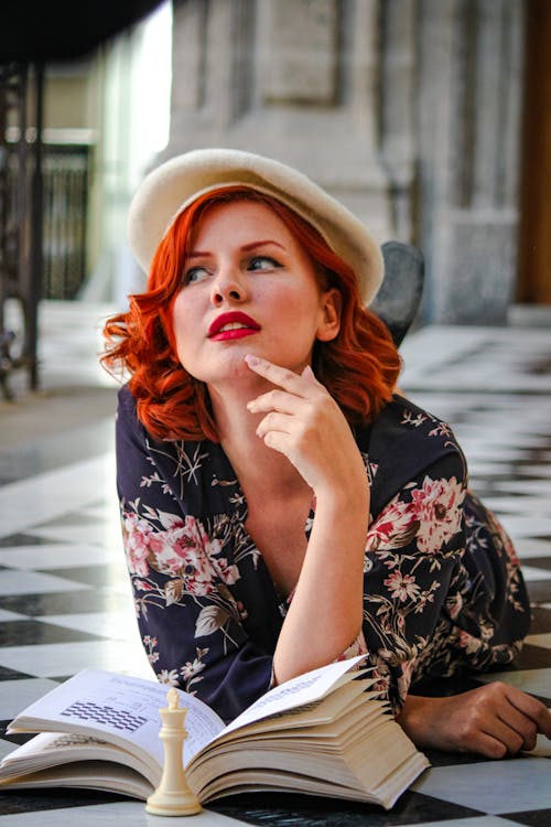 A woman with red hair and a hat is sitting on the floor with a book