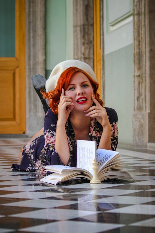 A woman with red hair and a hat is laying down on the floor reading a book
