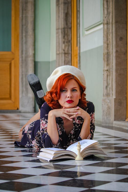 A woman with red hair and a hat is laying on the floor reading a book