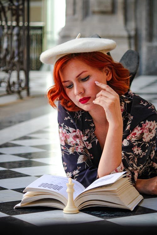 A woman with red hair and a white hat is reading a book