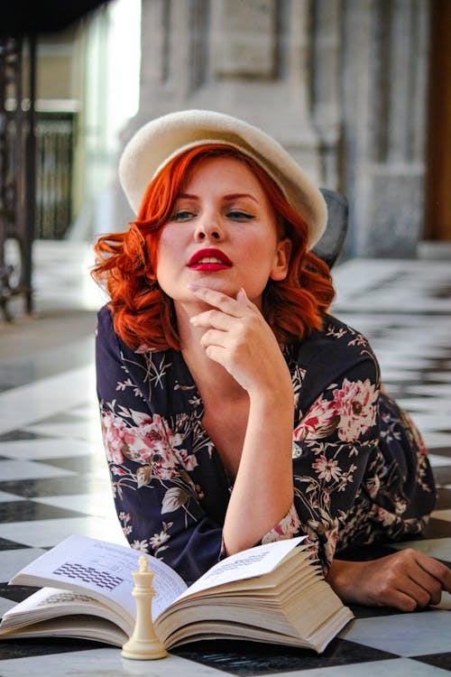 A woman with red hair and a hat is laying down on the floor