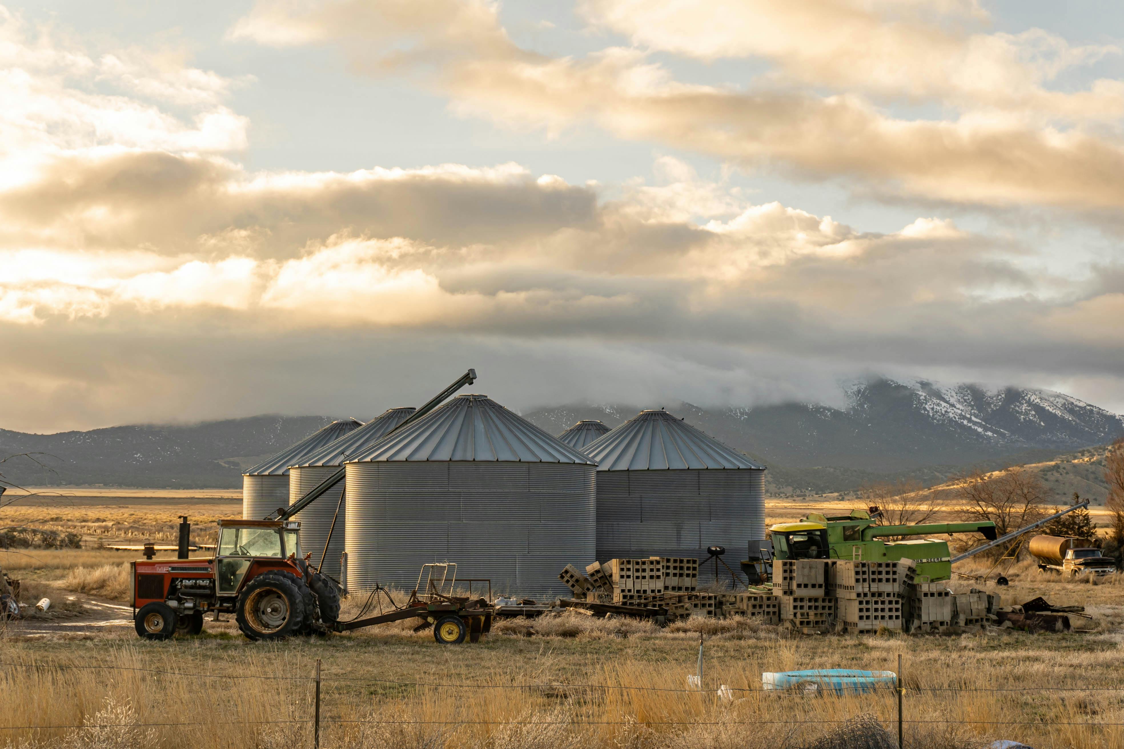 Serene rural landscape featuring silos, tractors, and a mountainous backdrop under cloudy skies.