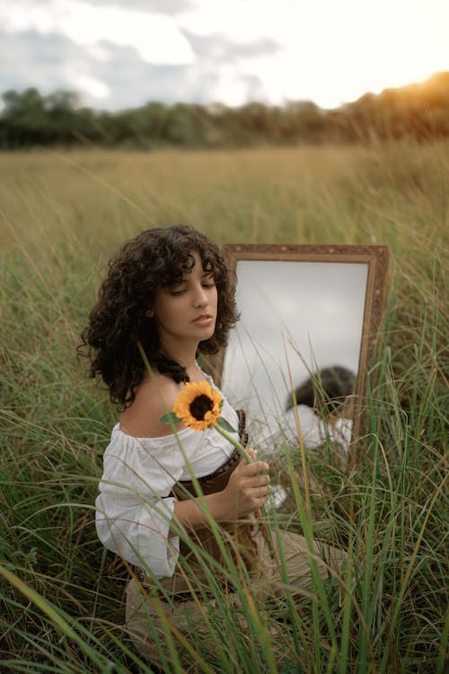 A woman in a field holding a sunflower and looking at a mirror