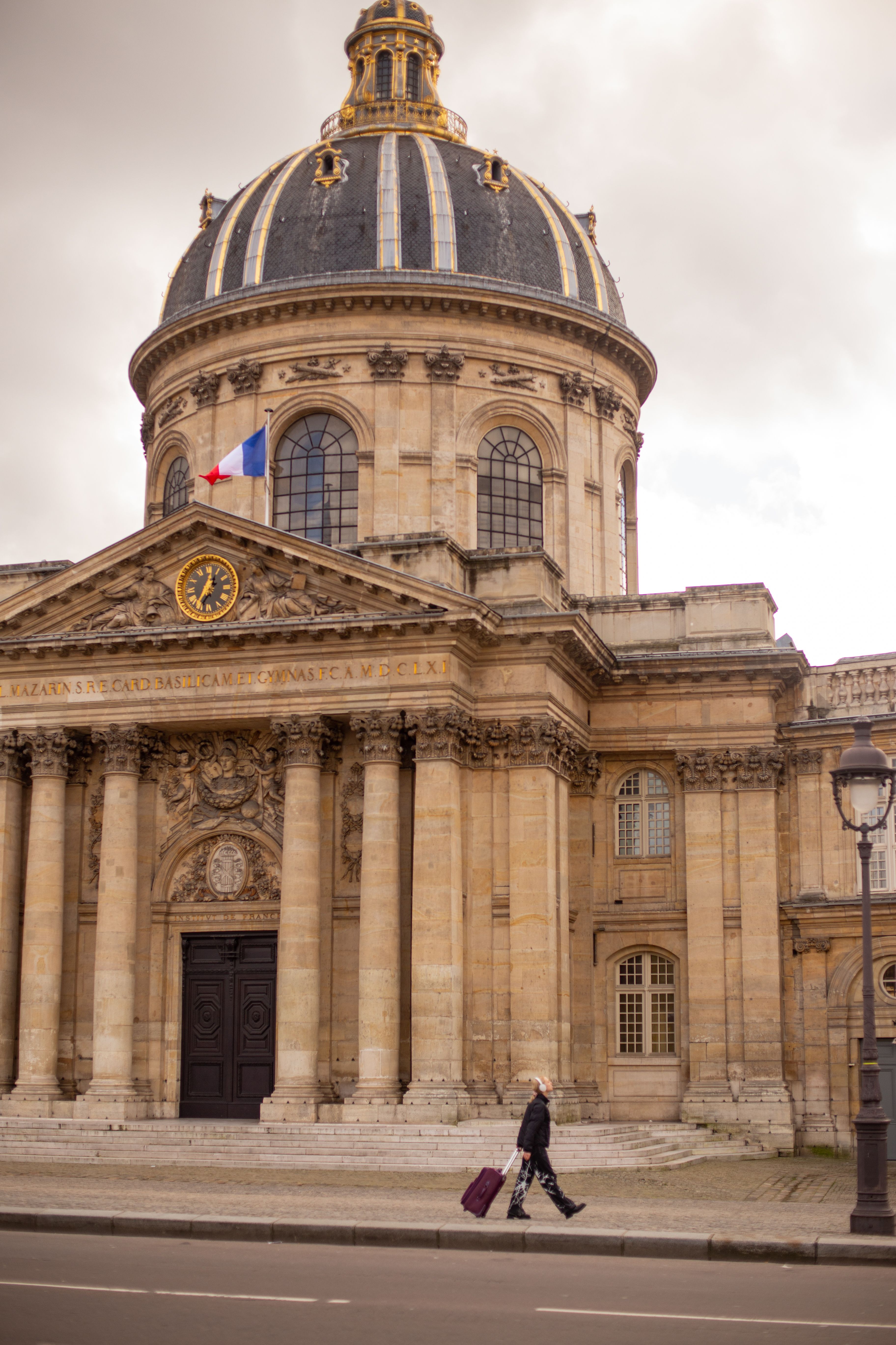 A person with a suitcase walks past the iconic Institut de France in Paris, highlighting classic French architecture.