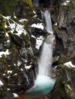 Serene winter cascade surrounded by snow-covered rocks in a lush mountainous area.