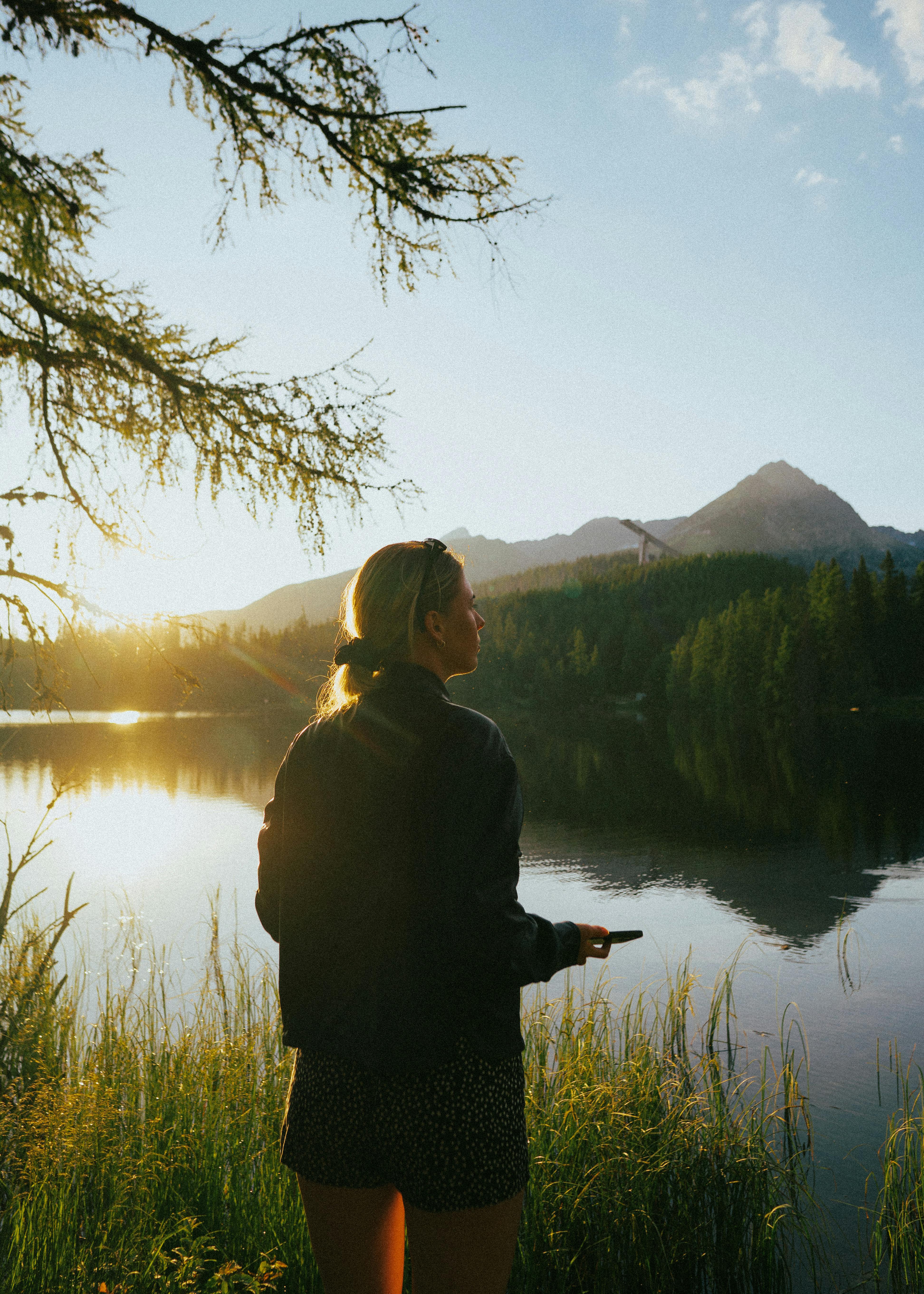 A woman stands by a serene lake with a mountain view, capturing the beauty of nature at sunset.