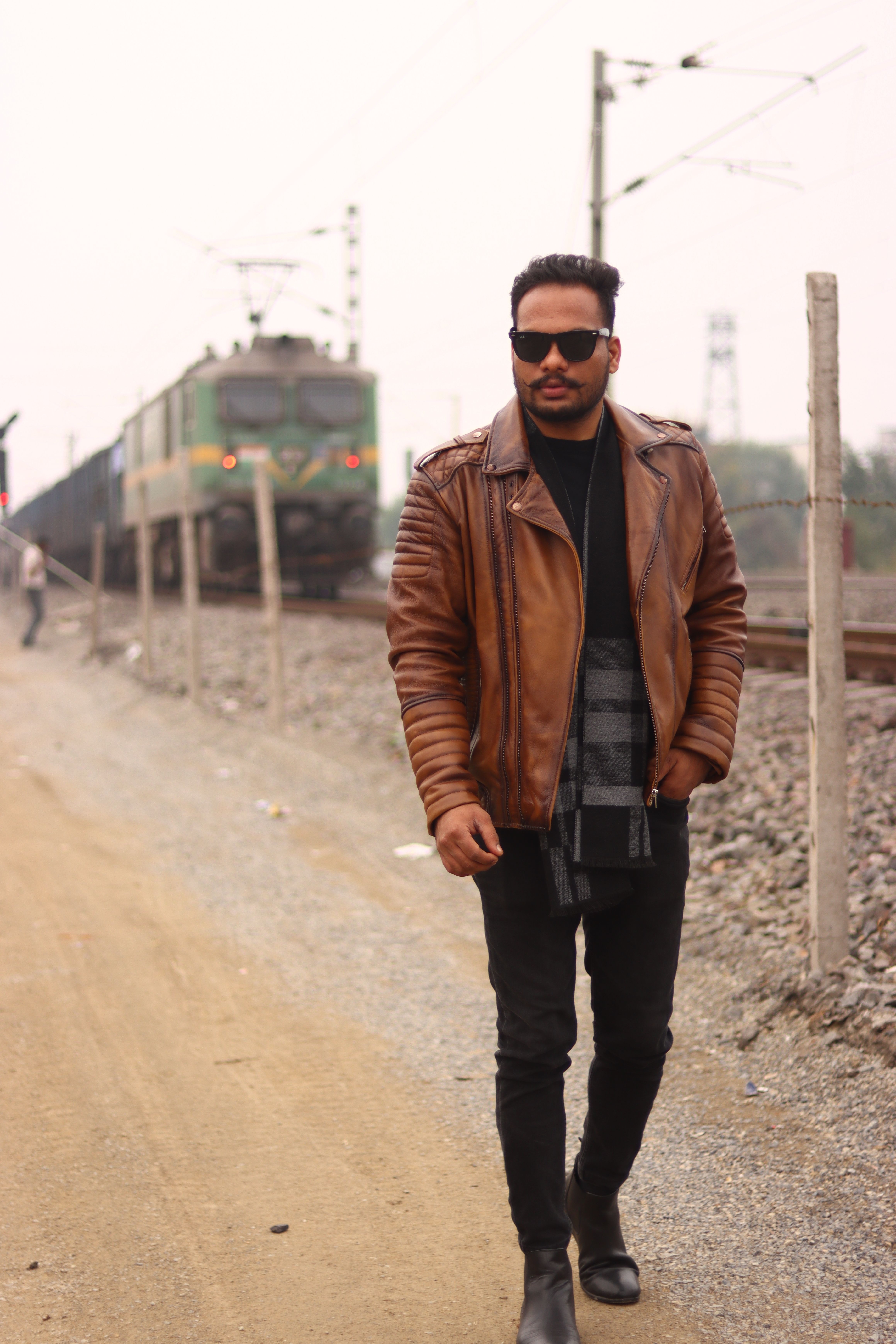 Man in a brown leather jacket walking by a train track, exuding urban style and confidence.
