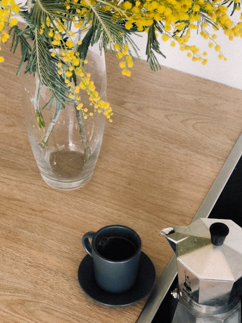 A coffee pot sits on a wooden counter with a vase of yellow flowers
