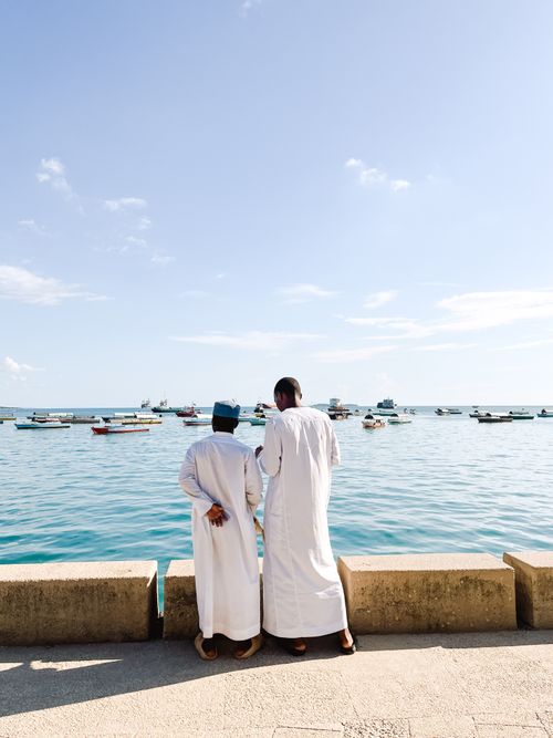 Free Two men in traditional attire stand by the waterfront in Zanzibar, with boats on the horizon. Stock Photo