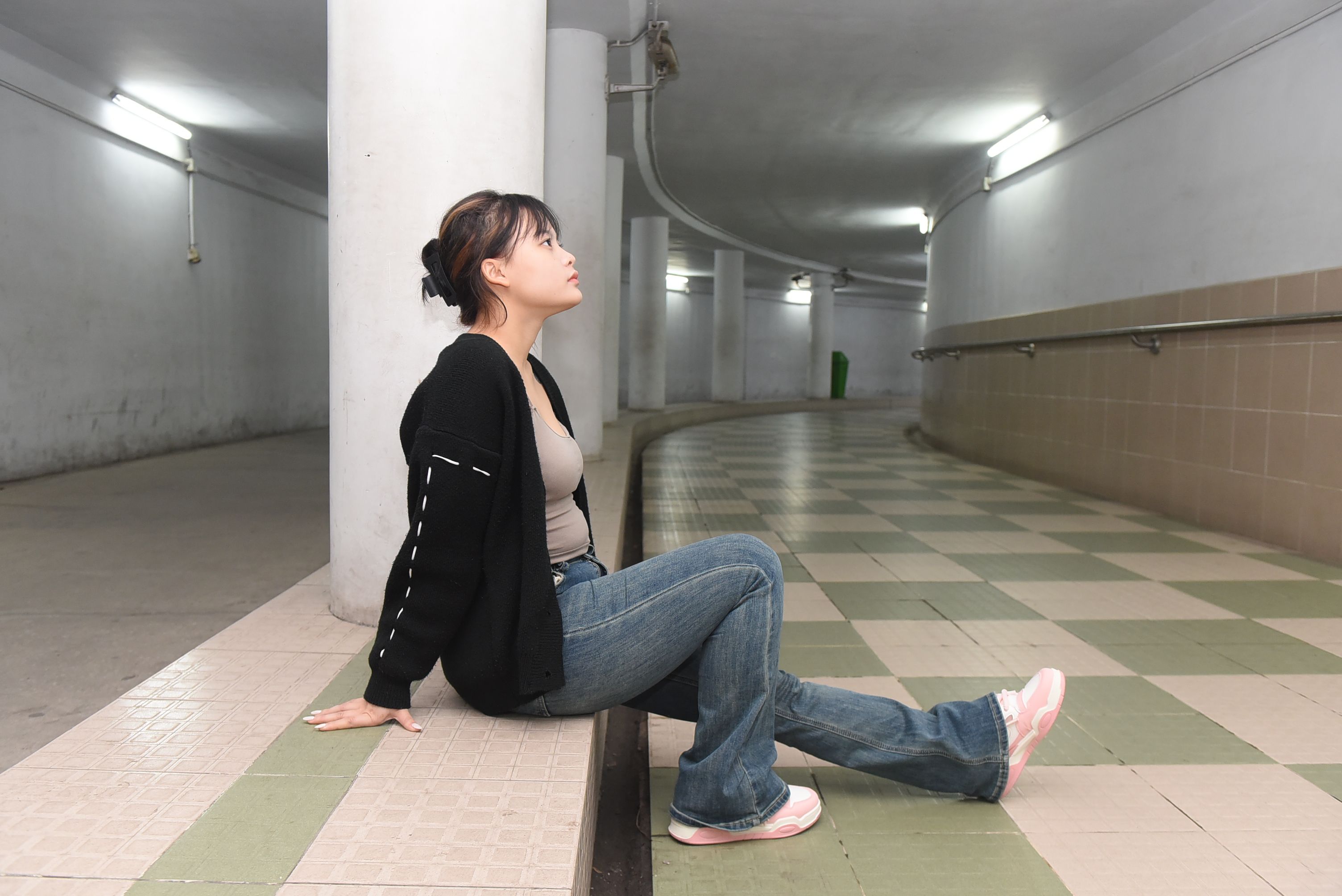 Young woman sitting in a tiled hallway, looking contemplative. Urban setting with modern interior.