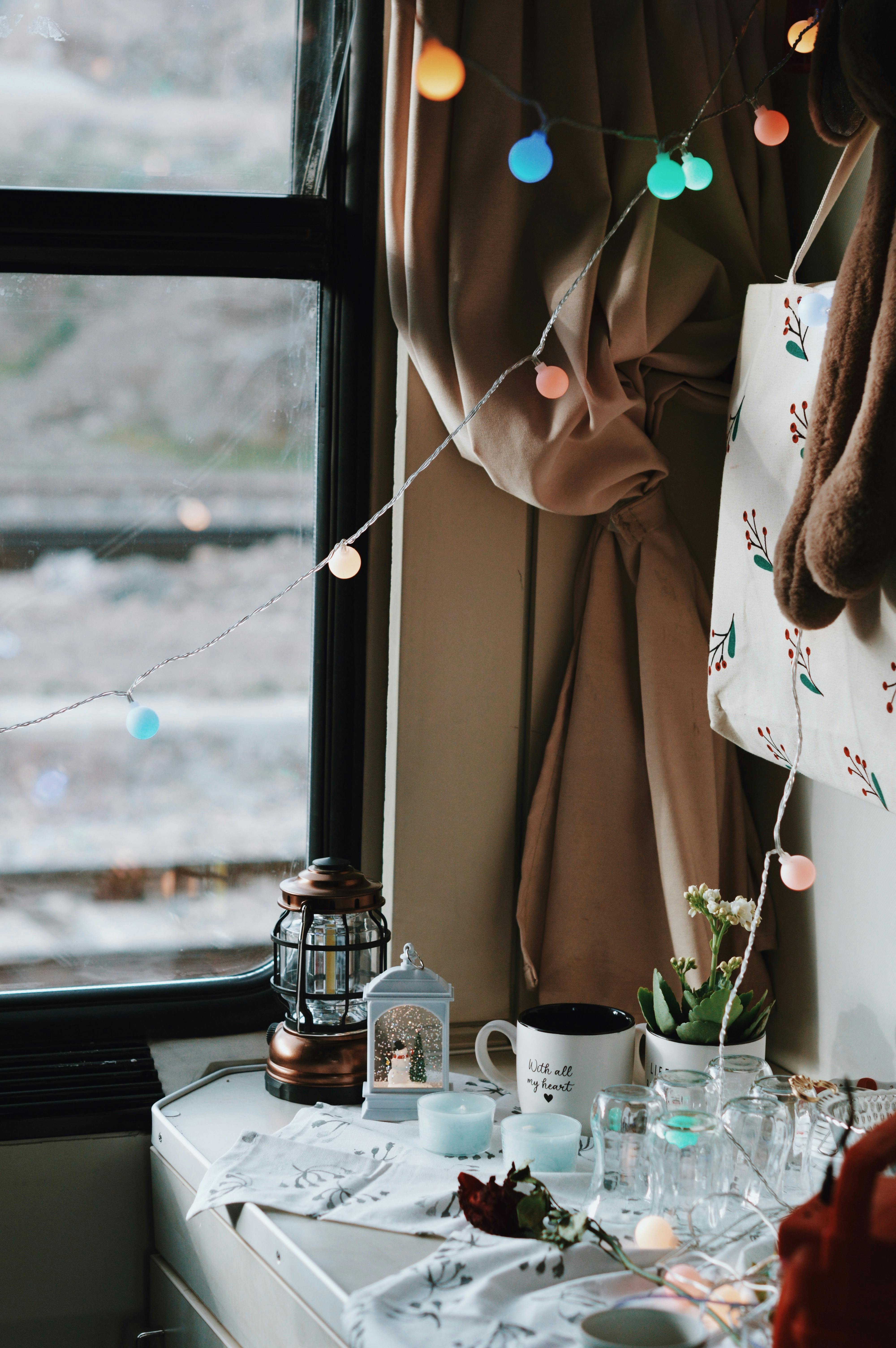 Warm and cozy indoor setup with string lights, lantern, and mugs on a windowsill.