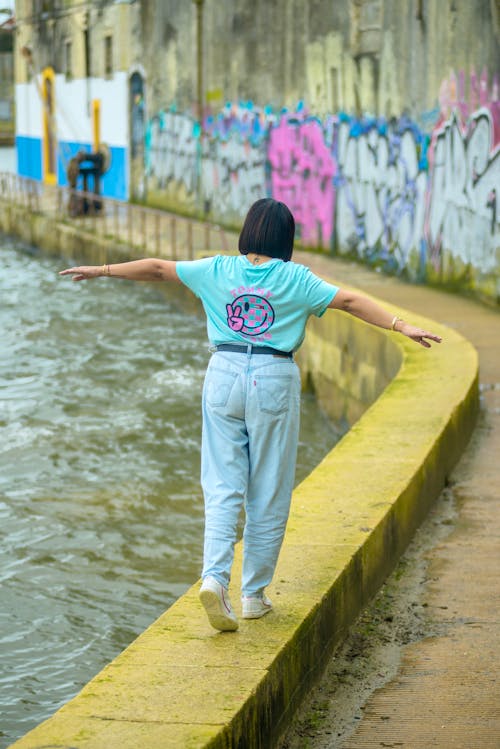 Free Woman in T-shirt and Jeans Walking with Arms Stretched on Wall over River Stock Photo