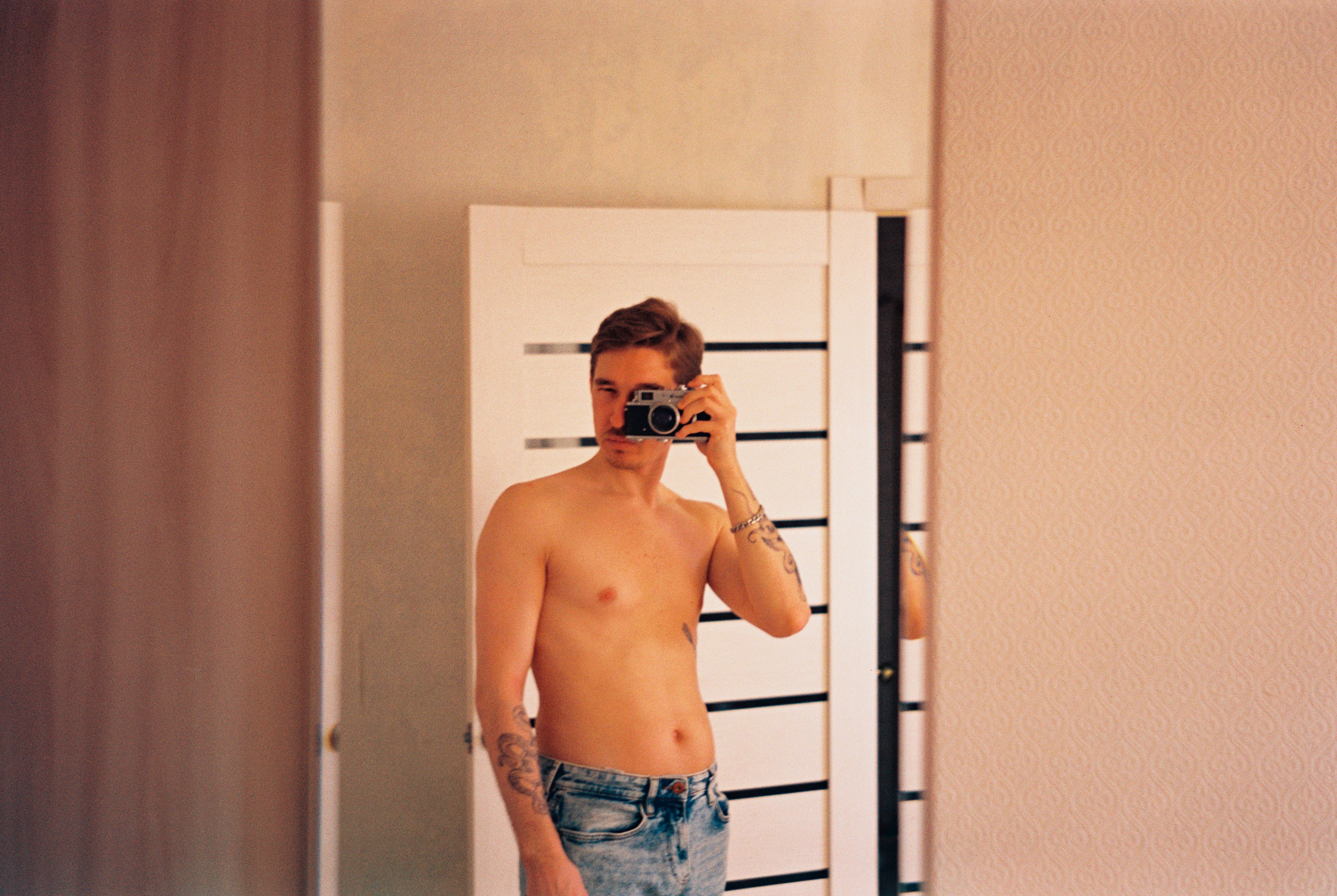 A man with tattoos takes a mirror selfie using an analog camera in an indoor setting.