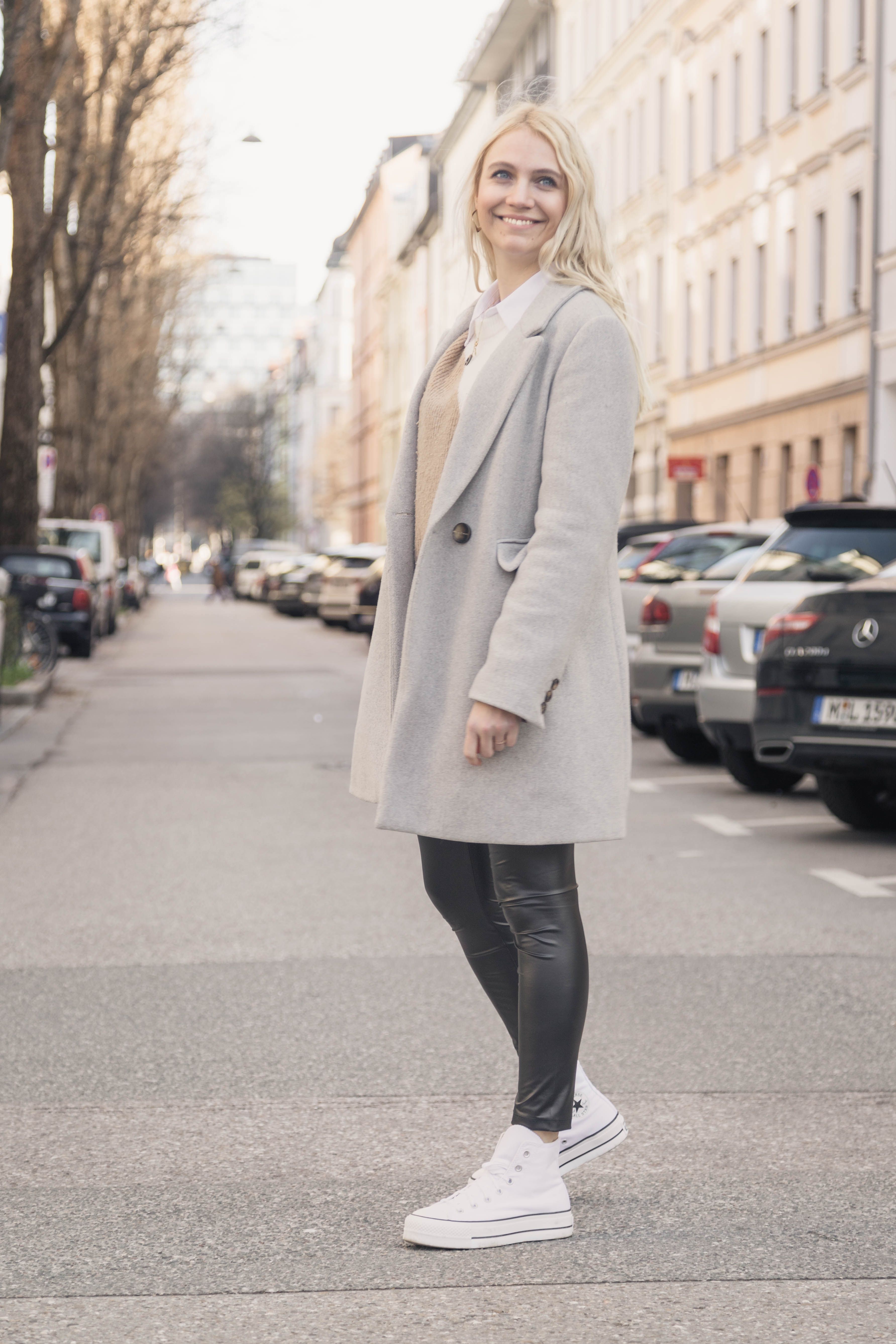Stylish young woman smiling in Munich city street wearing a gray coat, embracing urban fashion and lifestyle.