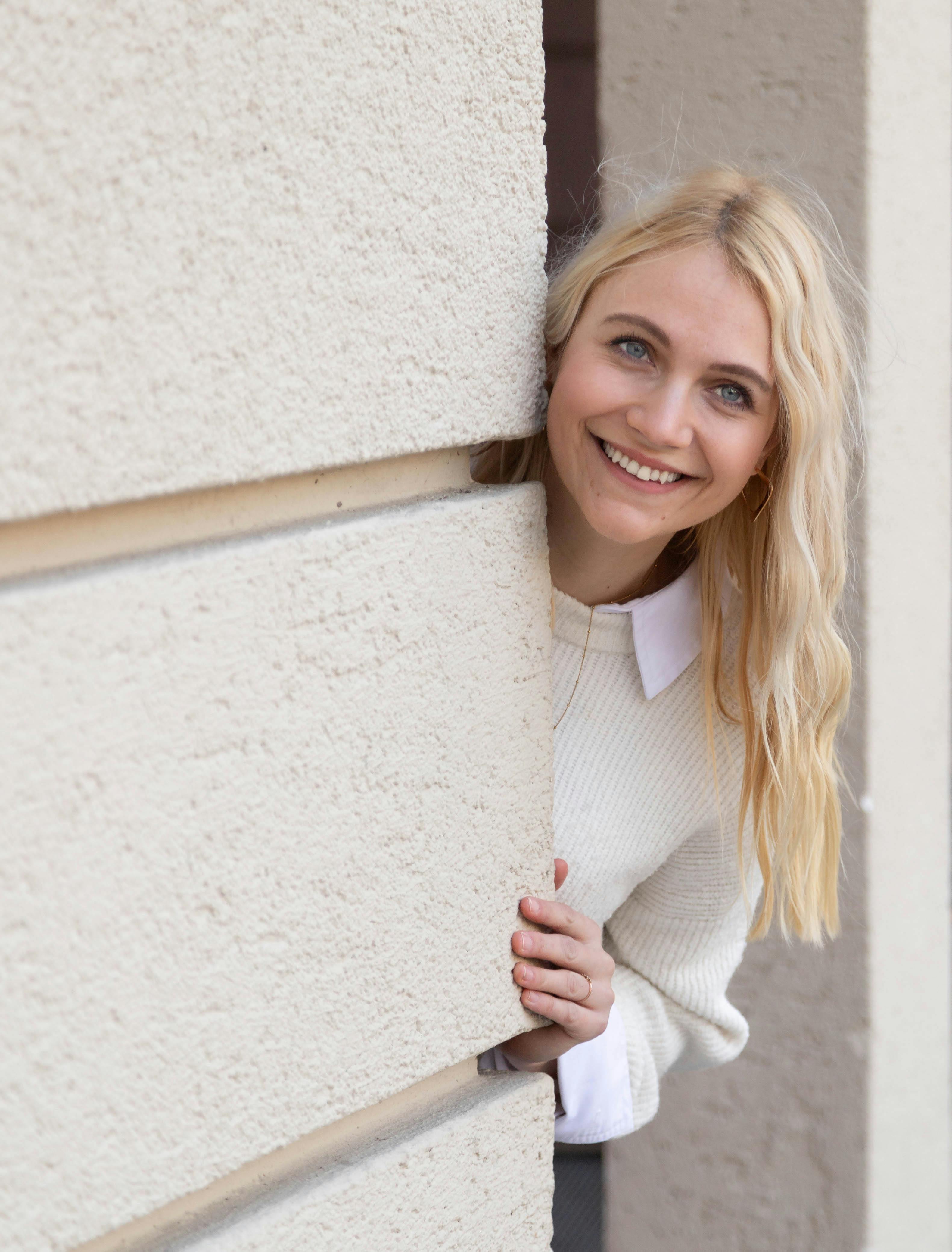 Blonde woman smiling and peeking around a wall in Munich. Casual and cheerful setting.