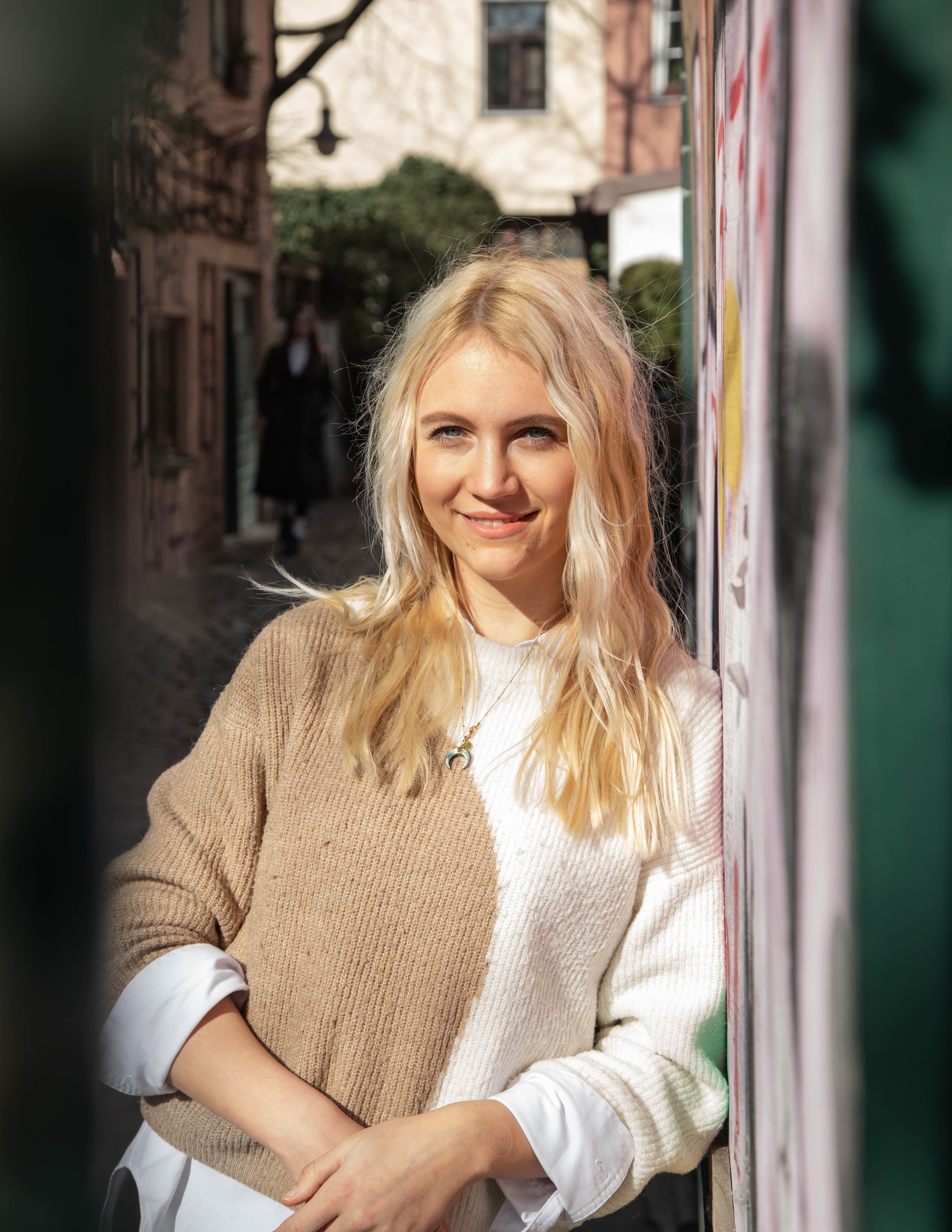 Portrait of a young blonde woman smiling in a sunny street in Munich, Germany, wearing a cozy sweater.